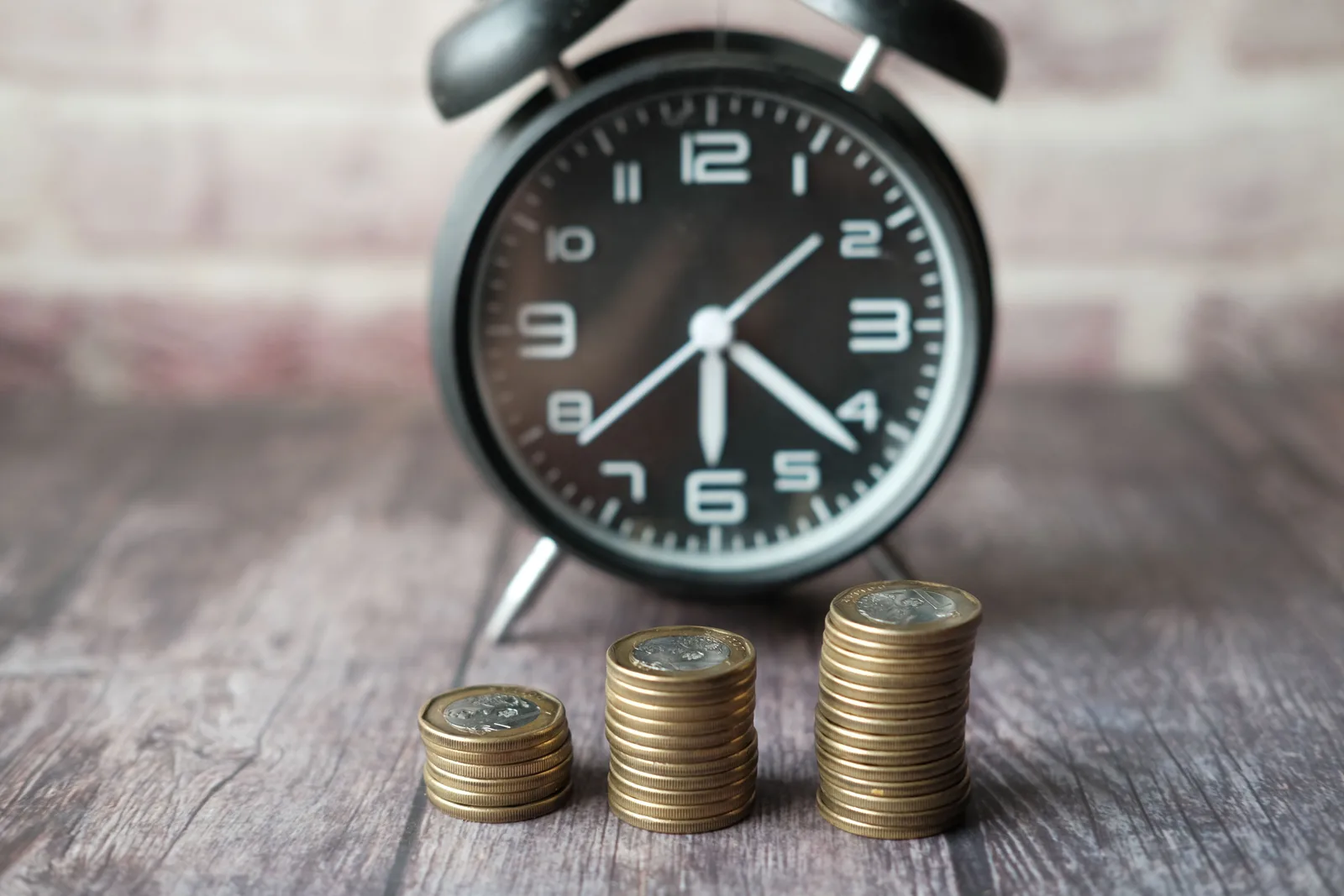 Reloj despertador junto a tres montones de monedas de tamaño creciente, como metáfora del interés compuesto y del paso del tiempo